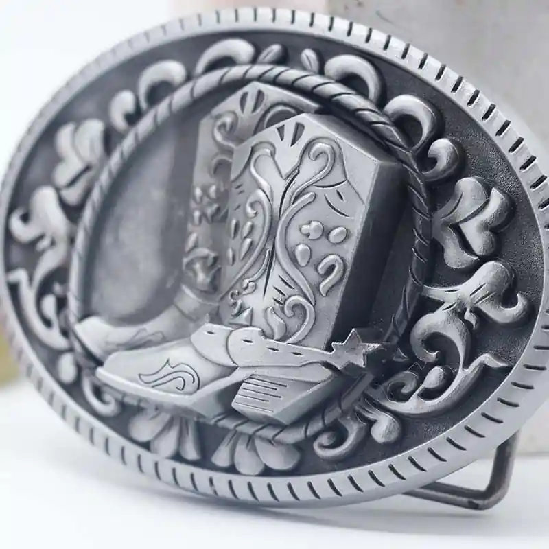 Close-up of a vintage western style metal belt buckle featuring embossed cowboy boots and ornate floral detailing in an antique silver finish.