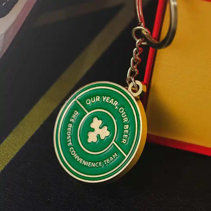 Close-up of a custom round enamel keychain with a green medal design, gold metal outline, and text “Our Year, Our Beer – DKS Bedste Convenience Team,” hanging from a metal chain.