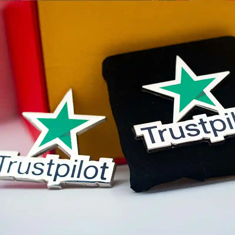 Two custom enamel pins featuring the Trustpilot green star logo and text, displayed on a colorful background with one pin placed inside a presentation box.