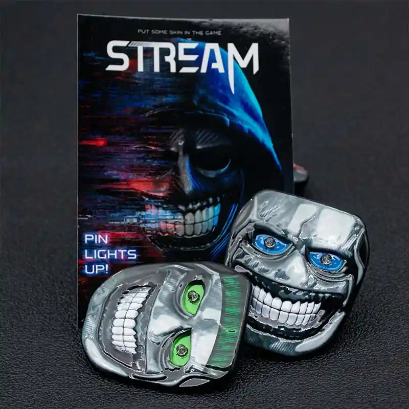Two metallic enamel pins featuring skull faces with colored LED eyes, displayed in front of a themed backing card on a dark surface.
