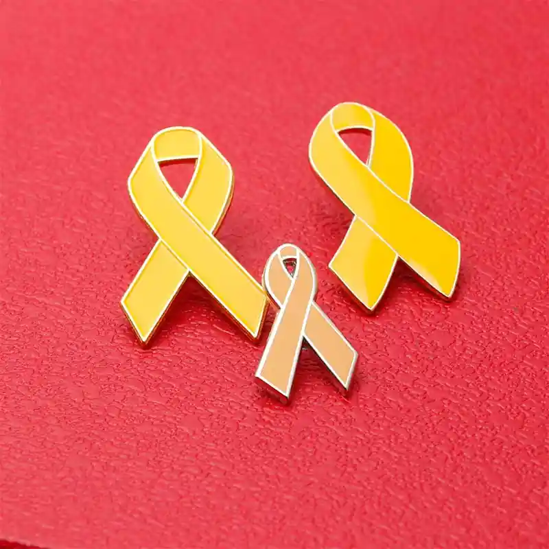 Three custom awareness ribbon enamel pins in yellow and gold tones displayed on a red textured background, representing support and awareness campaigns.