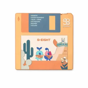 A retro floppy-disk–shaped PVC patch featuring the G-EIGHT logo, pastel desert scenery, and two cute cartoon characters, with text referencing gamers, indie games, and Taiwan, on a white background.