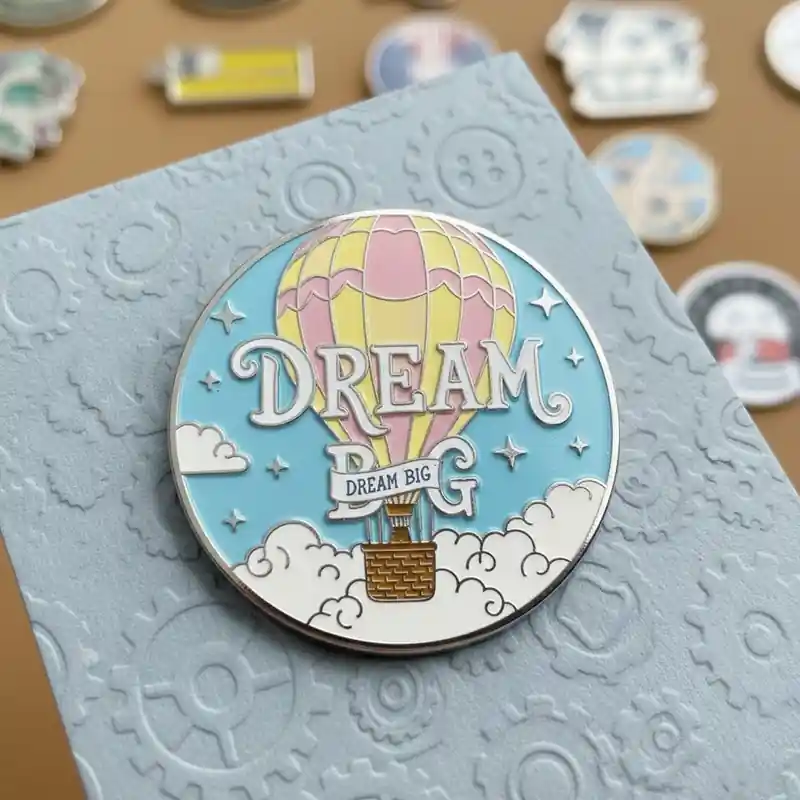 A pin featuring the phrase 'DREAM BIG' in bold lettering with a colorful hot air balloon floating among clouds, set against a light blue sky with stars, capturing a sense of adventure and hope.