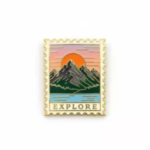 An enamel pin shaped like a postage stamp, featuring a colorful design of mountains, a sunset, and a body of water with the word "EXPLORE" at the bottom in gold.