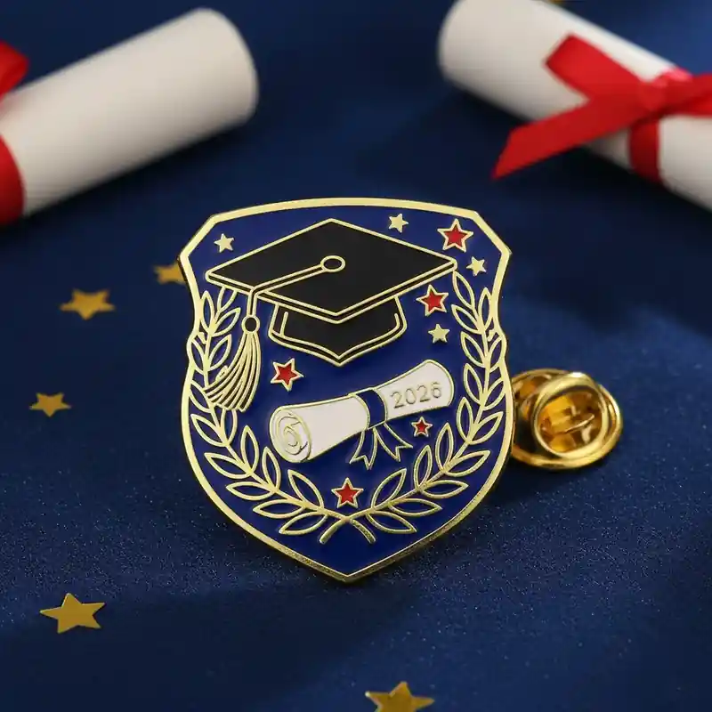 A gold-colored lapel pin shaped like a shield with a graduation cap, rolled diploma, and stars, commemorating the year 2026, set on a blue fabric background with scattered golden stars.