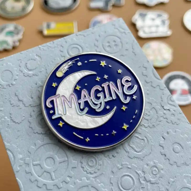 A pin featuring the word 'IMAGINE' in whimsical script, surrounded by a crescent moon and twinkling stars, set against a deep blue background, with a gear-themed gray card behind it.