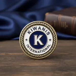 A circular Kiwanis pin featuring a bold blue and white design. The top part of the pin features the word "KIWANIS" in bold lettering, with a large letter "K" in the center. Surrounding the design is a gold rope-like border, and the word "INTERNATIONAL" appears below the "K." The pin represents the global efforts of Kiwanis International, a service organization dedicated to improving the lives of children.