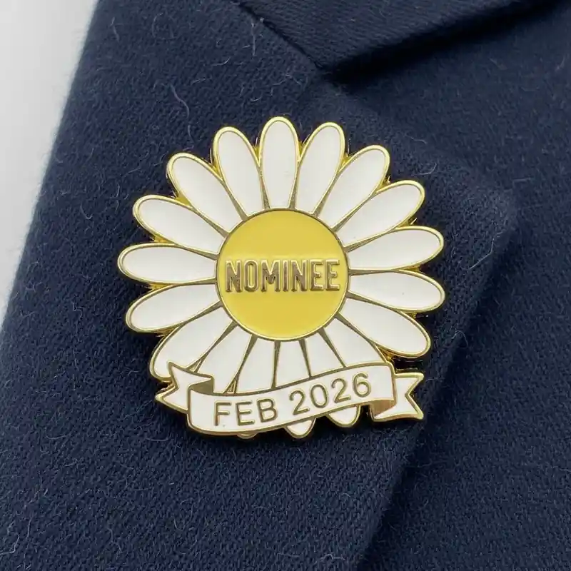 A yellow and white daisy pin with the text "NOMINEE" in the center and a ribbon at the bottom reading "FEB 2026."