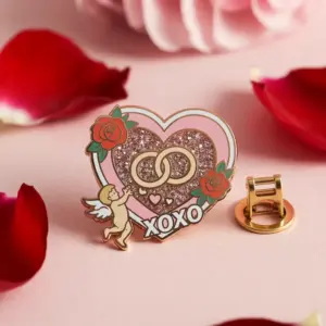 A heart-shaped enamel pin featuring glitter details, interlocking rings, roses, and a small cupid, with “XOXO” text on a soft pink background.
