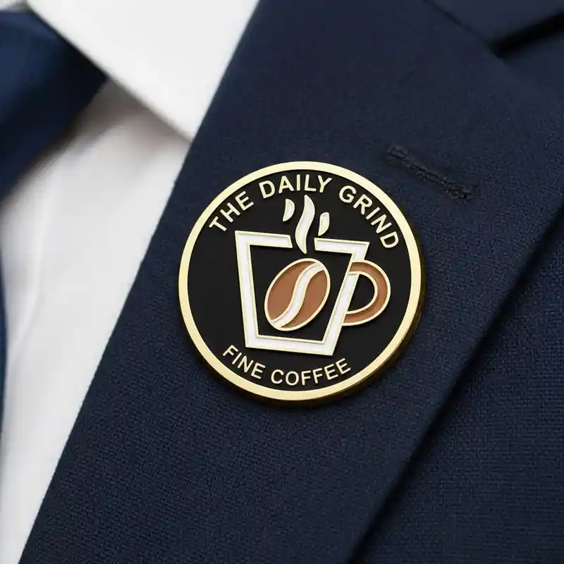 An enamel pin featuring a cup of coffee with steam rising, a coffee bean logo, and the text "THE DAILY GRIND" and "FINE COFFEE." The design is black and gold with a modern and clean aesthetic.