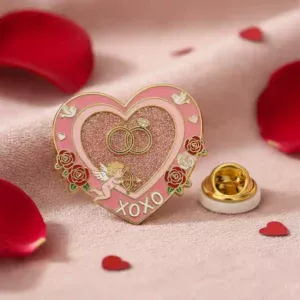 A pink heart-shaped lapel pin featuring two wedding rings, a cupid holding a bow and arrow, and the word "XOXO" with roses and birds, set on a soft pink fabric background.
