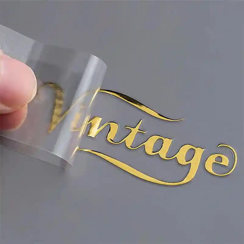 Image showing the process of peeling a clear backing off a gold metallic 3D transfer sticker with the word "Vintage" being revealed on a surface.