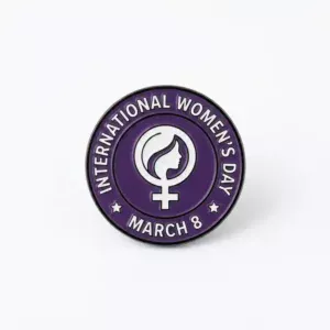 Round purple enamel pin featuring the female symbol with a woman silhouette and the text "International Women’s Day – March 8" around the border.