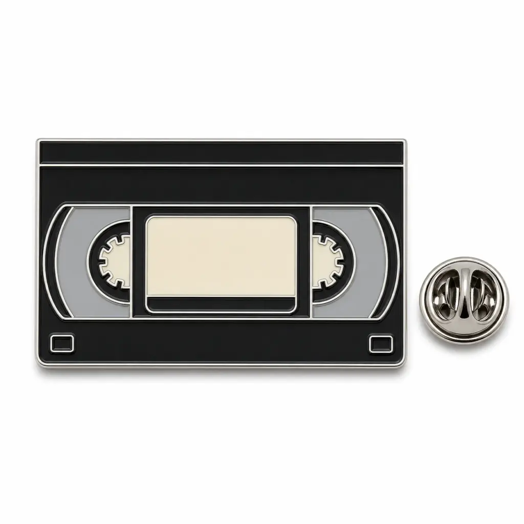 Badge en émail noir pour cassettes VHS avec étiquette crème vierge, bobines grises, intercalaires argentés et pochette papillon.