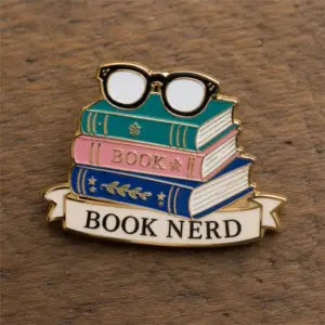 Book stack glasses Book Nerd enamel pin with gold metal outline, colorful stacked books, black glasses, white ribbon banner, Book Nerd lettering, and wood tabletop background.