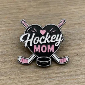 Heart sticks Hockey Mom enamel pin with silver metal outline, black heart background, crossed hockey sticks, pink accents, ice hockey puck, and Hockey Mom lettering on a wood tabletop background.