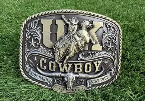 A custom western belt buckle featuring a cowboy riding a horse, raised lettering, star accents, and detailed antique metal engraving on a decorative silver and black finish.
