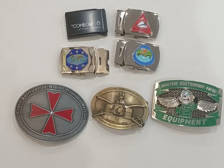 A collection of custom belt buckle samples displaying different metal finishes, enamel colors, embossed designs, and decorative craftsmanship details.