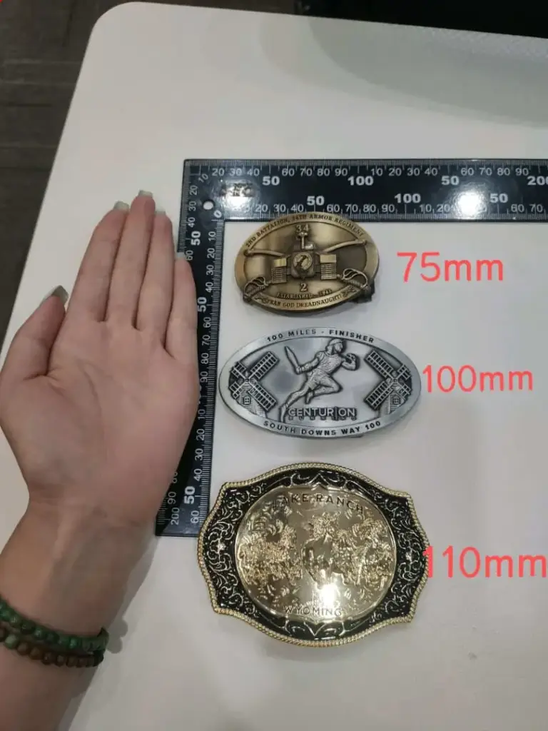 A size comparison guide showing custom belt buckles measured with a ruler, including buckle dimensions of 75mm, 100mm, and 110mm for different designs.